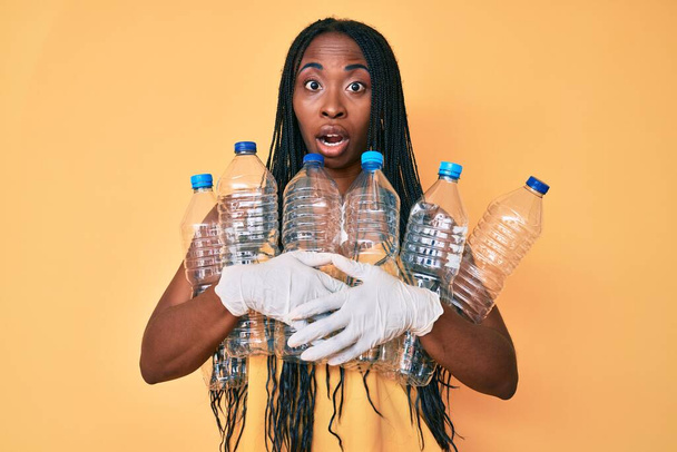 Femme afro-américaine avec des tresses tenant le recyclage des bouteilles en plastique dans le visage de choc, l'air sceptique et sarcastique, surpris avec la bouche ouverte  - Photo, image