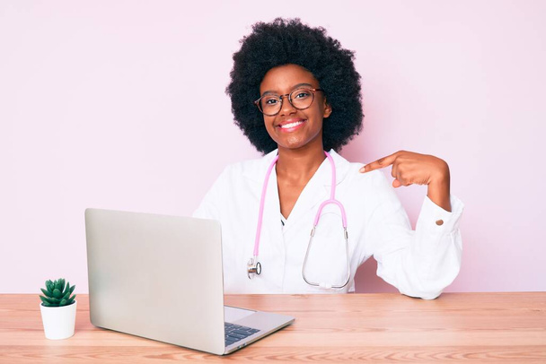 Молода афроамериканська жінка, одягнена в доктор стетоскоп, працює за допомогою комп'ютерного ноутбука, вказуючи пальцем на посмішку, щаслива і горда  - Фото, зображення