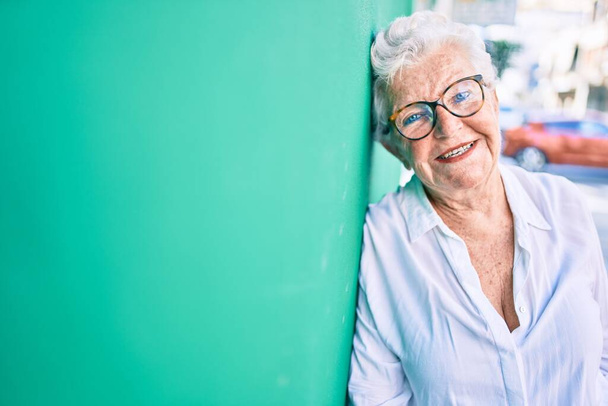 Ηλικιωμένη ηλικιωμένη γυναίκα με γκρίζα μαλλιά χαμογελαστή χαρούμενη ακουμπισμένη στον εξωτερικό τοίχο - Φωτογραφία, εικόνα