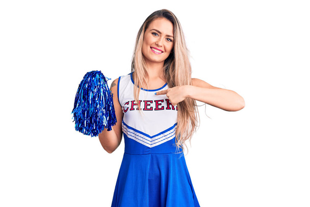 Young beautiful blonde woman wearing cheerleader uniform holding pompom pointing finger to one self smiling happy and proud  - Photo, Image