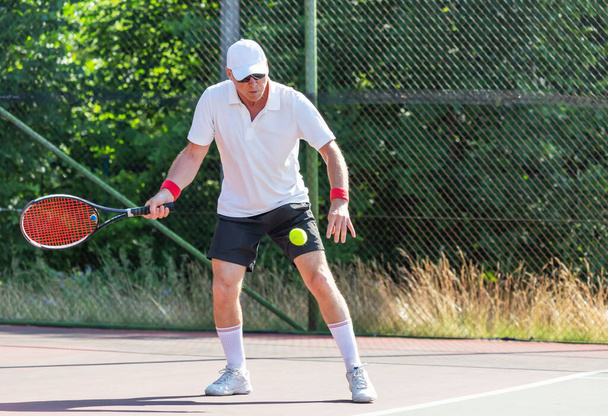 Un anciano en buena forma física juega al tenis en una cancha al aire libre en un día soleado - Foto, Imagen