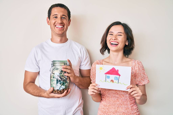 Красива пара тримає будинок малюнок і банку з доларами посміхається і сміється важко вголос, тому що смішний божевільний жарт
.  - Фото, зображення