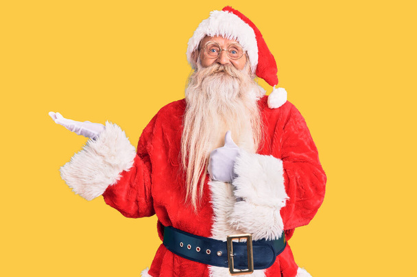 Viejo hombre mayor con el pelo gris y la barba larga con traje tradicional de Santa Claus mostrando la palma de la mano y haciendo buen gesto con los pulgares hacia arriba, sonriendo feliz y alegre  - Foto, imagen
