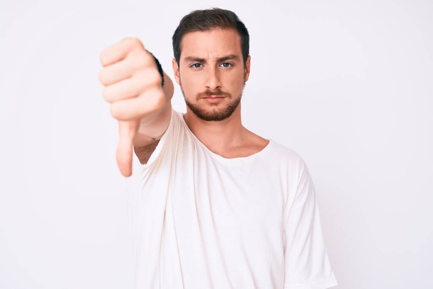 Young handsome man wearing casual white tshirt looking unhappy and angry showing rejection and negative with thumbs down gesture. bad expression.  - Fotoğraf, Görsel