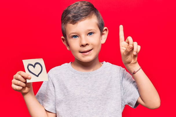 Cute blond kid holding heart reminder smiling with an idea or question pointing finger with happy face, number one  - Photo, image