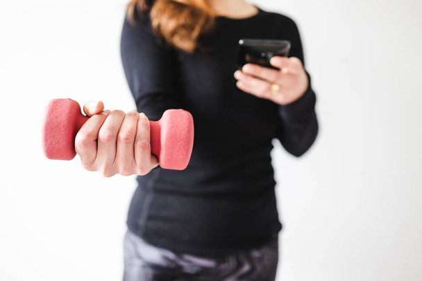 home fitness and online coaching concept, woman in sporty clothes holding dumbbell and smartphone to follow a fitness video or routine online - Foto, Imagen