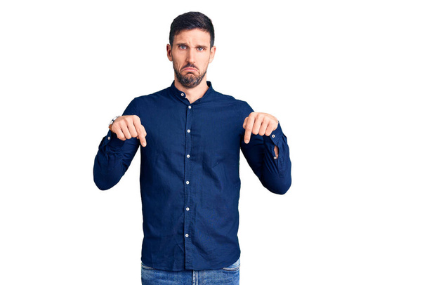 Jonge knappe man draagt casual shirt naar beneden wijzend droevig en overstuur, wijzend richting met vingers, ongelukkig en depressief.  - Foto, afbeelding