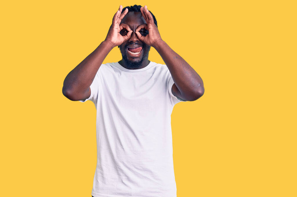 Jonge Afro-Amerikaanse man met vlechten in casual wit T-shirt doet ok gebaar als verrekijker steken tong uit, ogen kijken door vingers. gekke uitdrukking.  - Foto, afbeelding