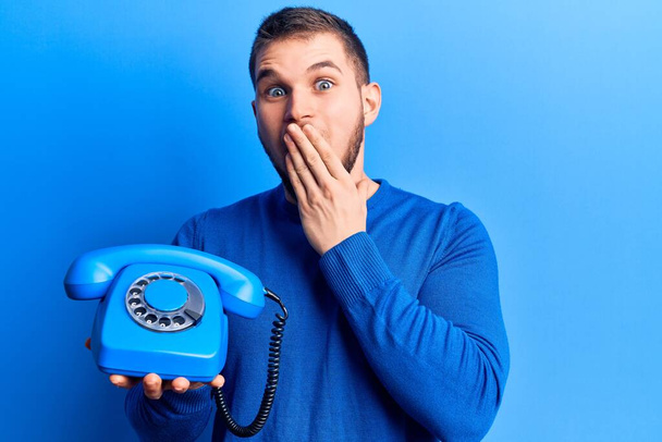 Homem bonito jovem segurando telefone vintage cobrindo a boca com a mão, chocado e com medo de erro. expressão surpreendida  - Foto, Imagem