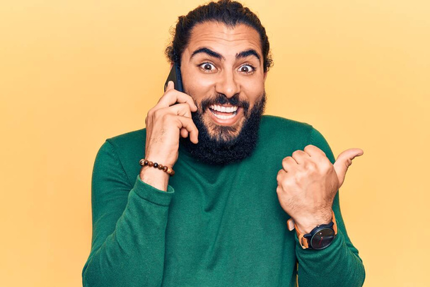 Hombre árabe joven teniendo conversación hablando en el teléfono inteligente apuntando pulgar hacia el lado sonriendo feliz con la boca abierta  - Foto, imagen