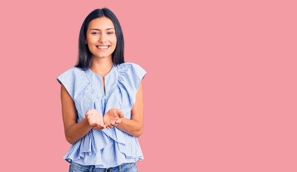 Νεαρή όμορφη Λατίνα κοπέλα φορώντας casual ρούχα χαμογελώντας με τα χέρια παλάμες μαζί λαμβάνοντας ή δίνοντας χειρονομία. αναμονή και προστασία  - Φωτογραφία, εικόνα