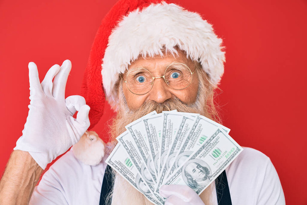 Staruszek z siwymi włosami i długą brodą w kostiumie Świętego Mikołaja trzymający dolary robiący znak ok palcami, uśmiechnięty przyjazny gest doskonały symbol  - Zdjęcie, obraz