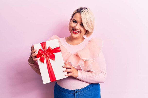 Young beautiful blonde plus size woman holding birthday gift over isolated pink background looking positive and happy standing and smiling with a confident smile showing teeth - Photo, Image