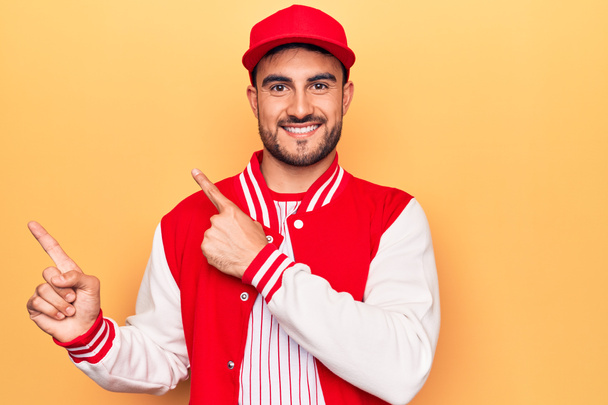 Hombre deportivo guapo con barba con chaqueta de béisbol y gorra sobre fondo amarillo sonriendo y mirando a la cámara apuntando con dos manos y dedos hacia un lado. - Foto, imagen
