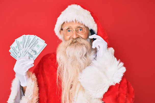 Viejo hombre mayor con traje de Santa Claus sosteniendo dólares y teléfono inteligente sonriendo con una sonrisa feliz y fresca en la cara. mostrando dientes.  - Foto, Imagen