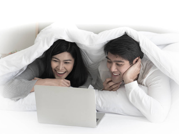 Aziatisch schattig jong paar liggen onder witte deken op hun bed genieten van het kijken naar film op notebook laptop in hun slaapkamer. Home entertainment concept - Foto, afbeelding