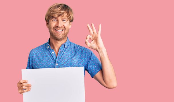 Handsome caucasian man with beard holding sale poster doing ok sign with fingers, smiling friendly gesturing excellent symbol  - Photo, Image