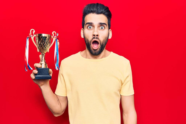Young handsome man with beard holding trophy scared and amazed with open mouth for surprise, disbelief face  - Photo, Image