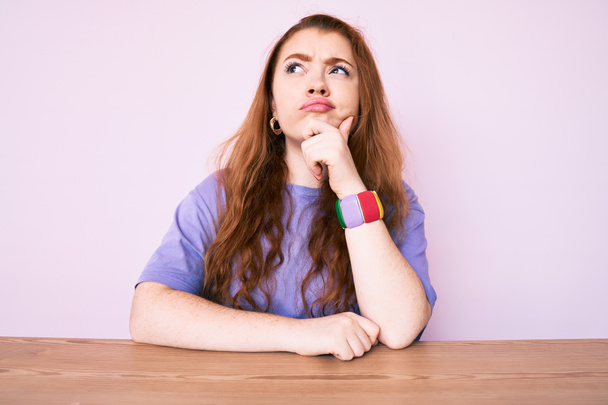 Young redhead woman wearing casual clothes sitting on the table thinking concentrated about doubt with finger on chin and looking up wondering  - Photo, Image
