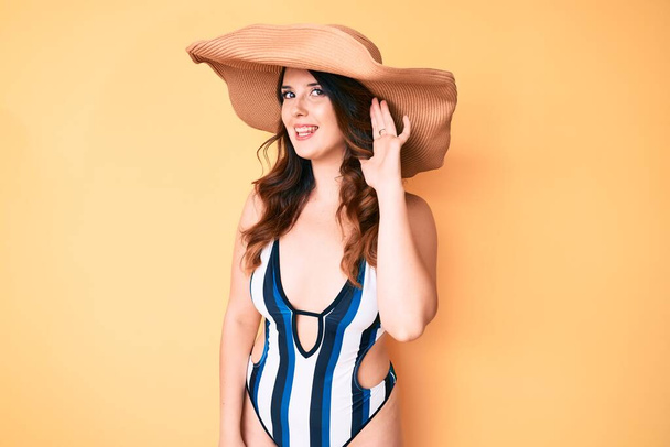 Young beautiful brunette woman wearing bikini and summer hat smiling with hand over ear listening and hearing to rumor or gossip. deafness concept.  - Photo, Image