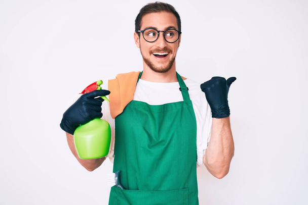 Young handsome man wearing apron holding sprayer pointing thumb up to the side smiling happy with open mouth  - Фото, зображення