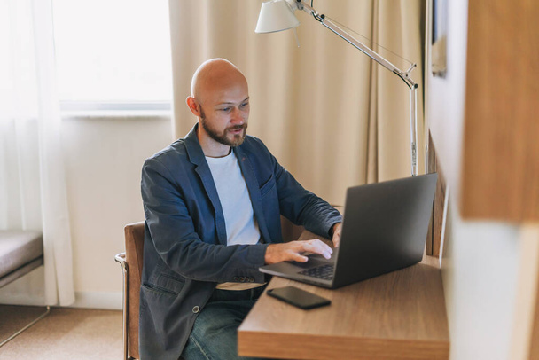 Homme barbu chauve adulte en veste bleue travaillant sur ordinateur portable dans la chambre d'hôtel - Photo, image