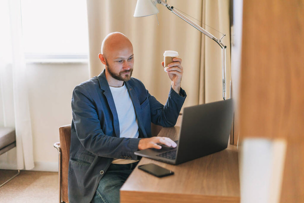 Adult bald bearded man in blue jacket with cup of coffee working on laptop in hotel room - Photo, Image
