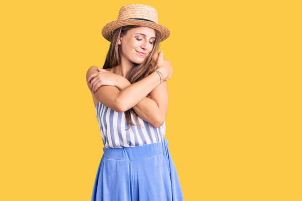 Jovem mulher loira bonita usando chapéu de verão abraçando-se feliz e positivo, sorrindo confiante. auto-amor e auto-cuidado  - Foto, Imagem