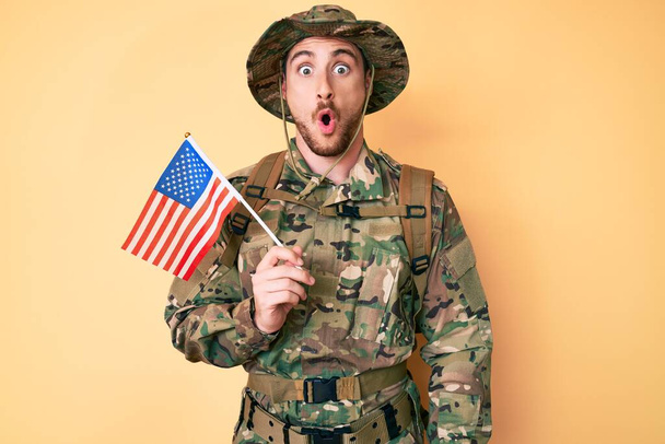 Giovane uomo caucasico con indosso camuffamento uniforme dell'esercito con bandiera USA spaventato e stupito a bocca aperta per la sorpresa, faccia incredulo  - Foto, immagini