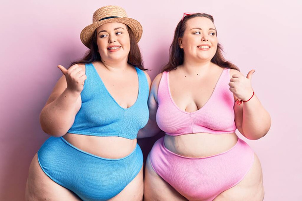 Young plus size twins wearing bikini smiling with happy face looking and pointing to the side with thumb up.  - Photo, Image