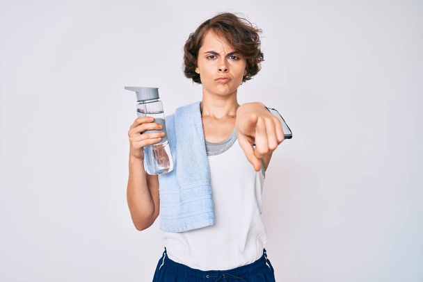 Junge hispanische Frau in Sportbekleidung und Handtuch, die eine Flasche Wasser trinkt und mit dem Finger in die Kamera und zu dir zeigt, selbstbewusste Geste, die ernst aussieht  - Foto, Bild