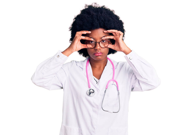 Young african american woman wearing doctor coat and stethoscope trying to open eyes with fingers, sleepy and tired for morning fatigue  - Foto, Imagen