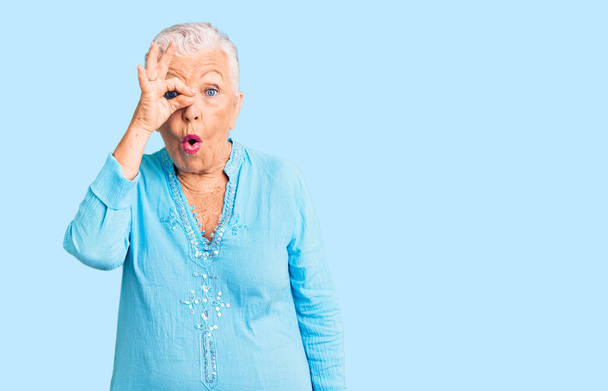 Старша красива жінка з блакитними очима і сірим волоссям носить літнє плаття, роблячи нормальний жест шокованим здивованим обличчям, дивиться через пальці. невіруючий вираз
.  - Фото, зображення