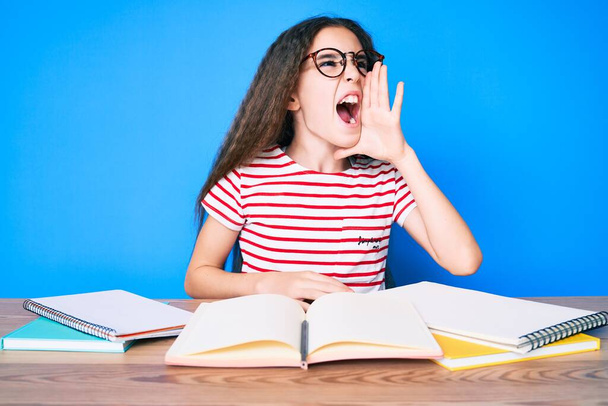 学校の試験のために勉強しているかわいいヒスパニック系の子供の女の子は、大声で叫び、口の上に手で横に大声で叫んでテーブルの上に座っている。コミュニケーションの概念.  - 写真・画像