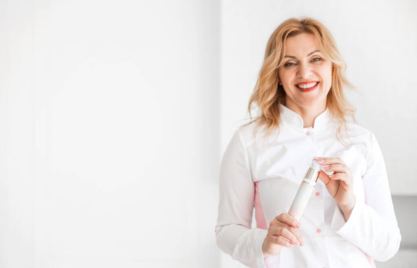 Стильная и красивая женщина-косметолог в белом медицинском костюме улыбается и смотрит в камеру. Женщина держит в руках косметический продукт. Горизонтальное фото - Фото, изображение