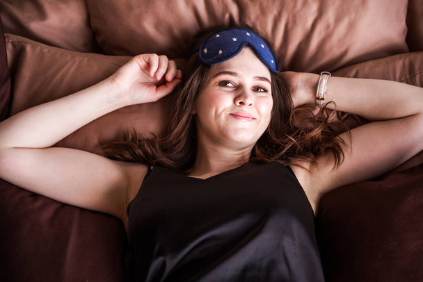 Na posteli leží roztomilá dívka v černém pyžamu a s maskou na spaní a dívá se do kamery. Pohled shora. Vodorovná fotografie - Fotografie, Obrázek
