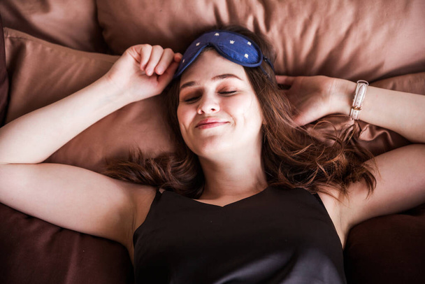 Siyah pijamalı ve yüzünde uyku maskesi olan güzel esmer bir kadın gözleri kapalı yatıyor. Yatay fotoğraf - Fotoğraf, Görsel