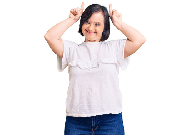 Down sendromlu esmer bir kadın, günlük beyaz tişört giyiyor ve boynuzlarını parmağında oynatarak komik jestler yapıyor.  - Fotoğraf, Görsel
