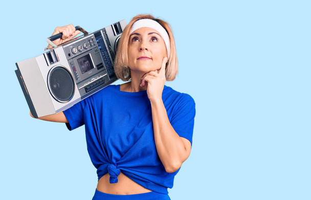 Young blonde woman wearing sportswear holding boombox, listening to music serious face thinking about question with hand on chin, thoughtful about confusing idea  - Фото, изображение