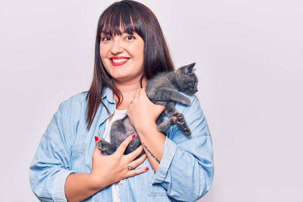 Young beautiful plus size woman smilling happy and confident. Standing with smile on face holding adorable cat over isolated white background - Foto, afbeelding