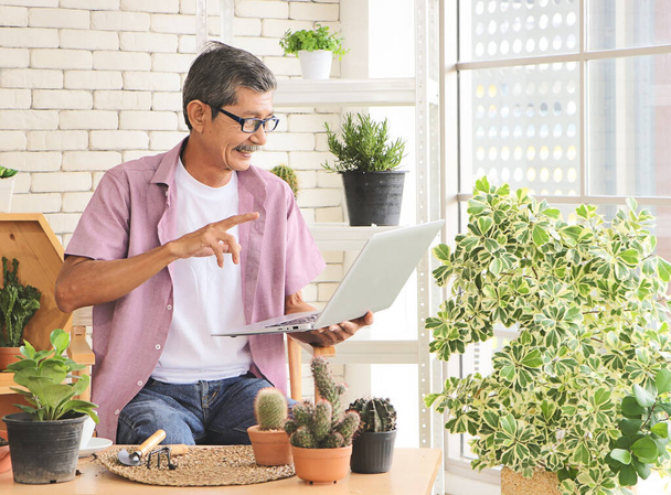 Senior people lifestyle and technology  concept. Happy Asian elderly man sitting at table with houseplants and gardening tools , making video call and smiling on laptop. - Photo, Image