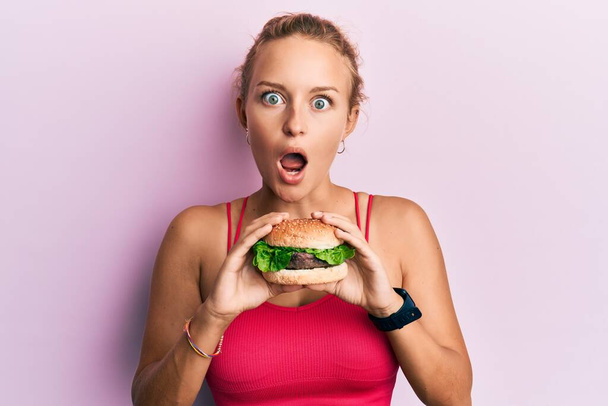 Beautiful caucasian woman eating a tasty classic burger afraid and shocked with surprise and amazed expression, fear and excited face.  - Valokuva, kuva