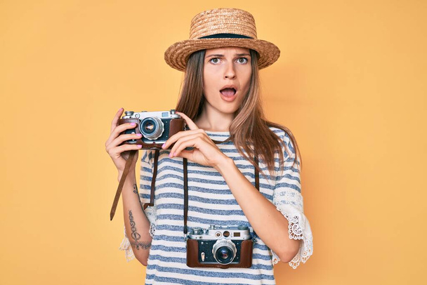 Beautiful caucasian tourist woman holding vintage camera in shock face, looking skeptical and sarcastic, surprised with open mouth  - Fotó, kép