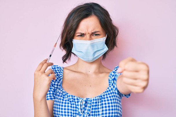 Young beautiful woman wearing medical mask holding syringe annoyed and frustrated shouting with anger, yelling crazy with anger and hand raised  - Photo, Image