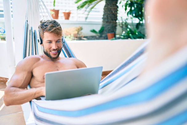 Jóképű fehér férfi mosolyog boldogan pihen egy függőágyon a teraszon, otthonról dolgozik, call center headsetet visel. - Fotó, kép