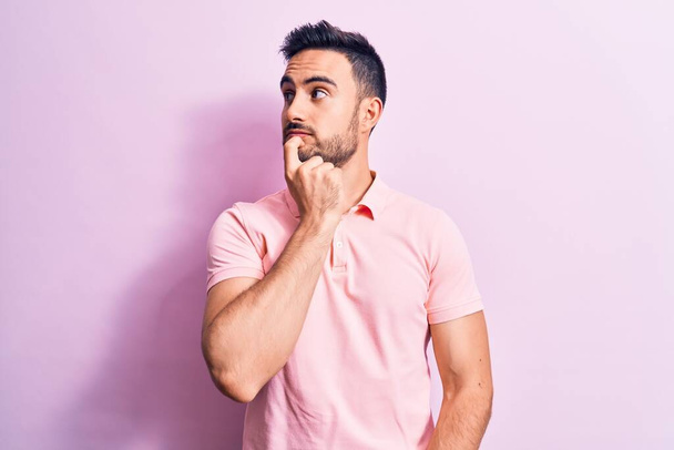 Young handsome man with beard wearing casual polo standing over pink background thinking concentrated about doubt with finger on chin and looking up wondering - Valokuva, kuva