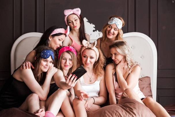 Muitas meninas alegres, brilhantes, positivas em pijama e com headbands cosméticos encenaram uma festa de pijama. As raparigas são fotografadas juntas ao telefone. Fotografia horizontal - Foto, Imagem