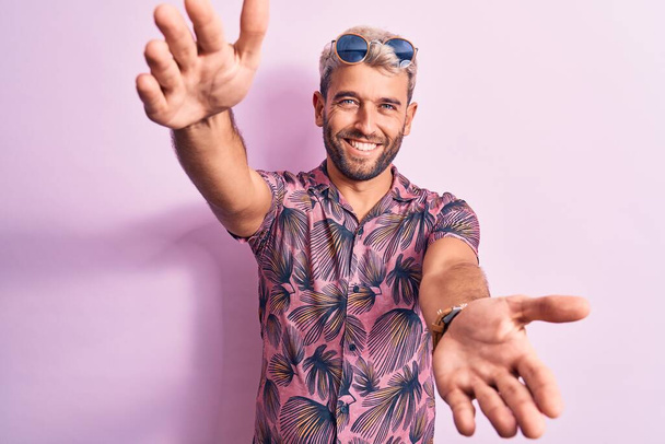 Knappe blonde man op vakantie met casual shirt en zonnebril over roze achtergrond kijkend naar de camera glimlachend met open armen voor knuffel. Vrolijke uitdrukking die geluk omarmt. - Foto, afbeelding