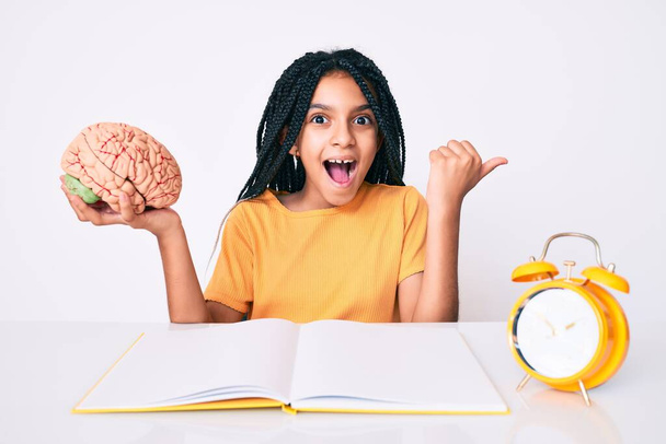 Jovem menina afro-americana criança com tranças segurando o cérebro enquanto estudava para a escola apontando o polegar para o lado sorrindo feliz com a boca aberta  - Foto, Imagem