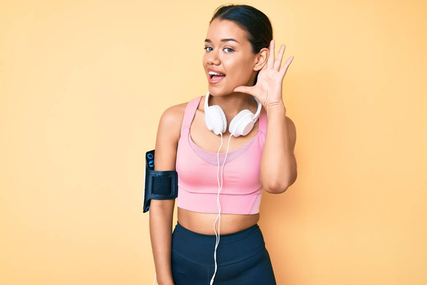 Молодая красивая латинская девушка в спортивной одежде и в наушниках, улыбаясь рукой над ухом, слушая и слушая слухи или сплетни. концепция глухоты.  - Фото, изображение